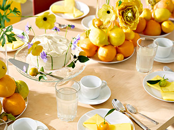 Mesa luminosa decorada en tonos amarillos con una tarta blanca, cítricos frescos, flores y vajilla blanca para una celebración alegre.