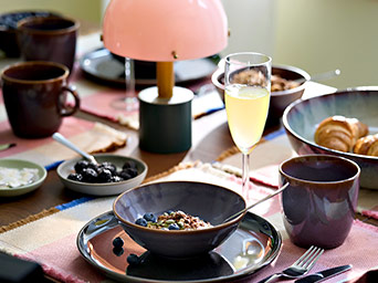 Mesa de desayuno con vajilla en tonos oscuros, boles con fruta y cereales, una copa de zumo y una pequeña lámpara rosa de diseño suave.