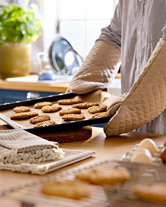 Persona con guantes de horno beige saca una bandeja de galletas recién horneadas sobre una mesa de madera con ingredientes.