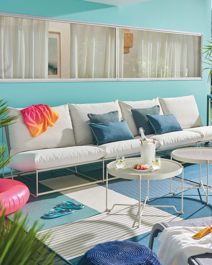 Modern terrace with a white sofa and blue cushions, round table with drinks and fruit, summer decor featuring a pink float and sandals on a blue rug.