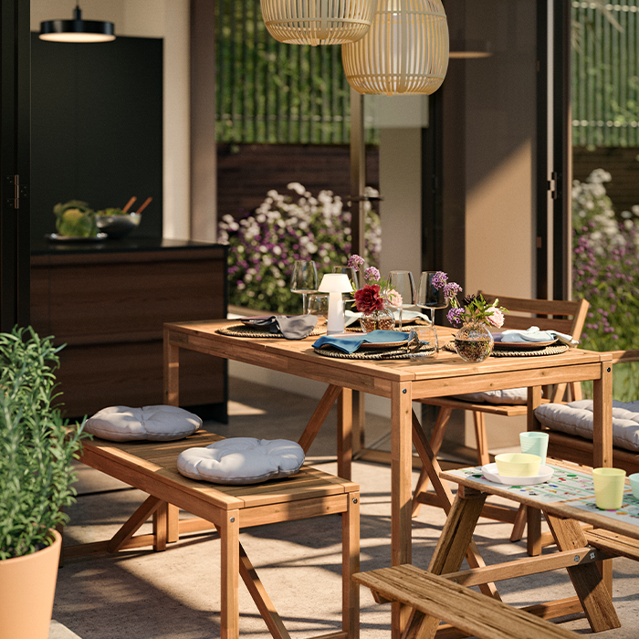 Sun-dappled patio with a wooden dining table set with wildflowers, glassware, and a view of a modern kitchen through glass doors.