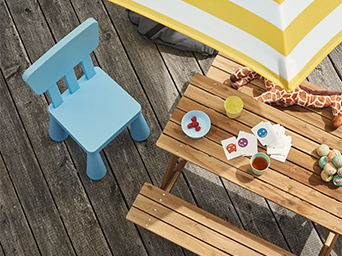 Top-down view of a wooden children's picnic table with snacks and toys, and a separate blue plastic chair, on a sunlit deck.
