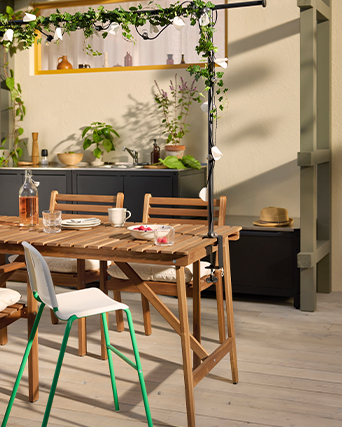 Wooden outdoor dining table set with drinks, snacks, and a straw hat, under a decorative ivy garland in warm sunlight.