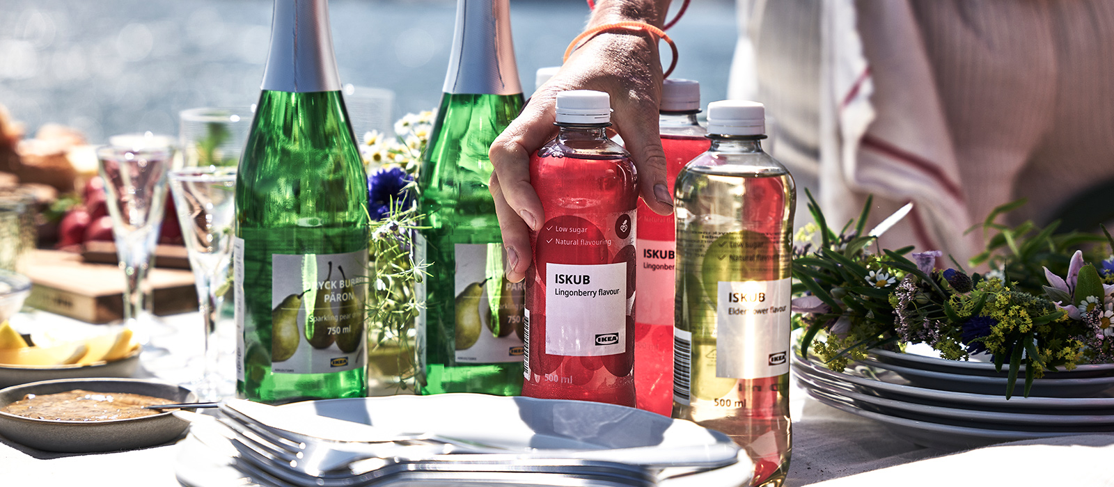 Outdoor table by the sea with bottles of ISKUB lingonberry drink and pear cider. A hand reaches for a red bottle near stacked white plates.