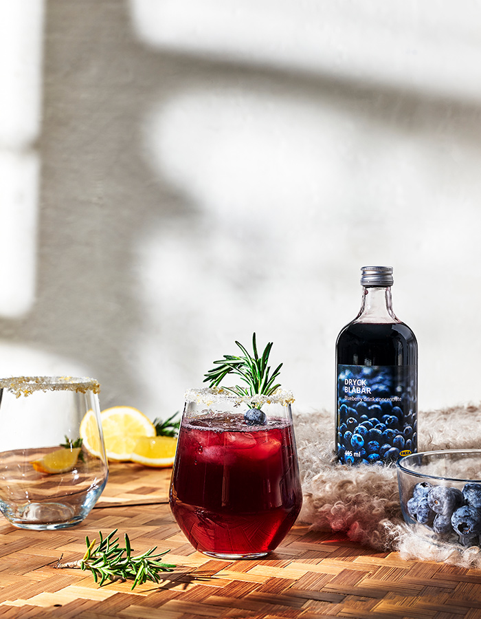 A glass of blueberry drink with ice and a rosemary sprig on a wooden table, next to a bottle of DRYCK BLÅBÄR concentrate and a bowl of fresh blueberries.