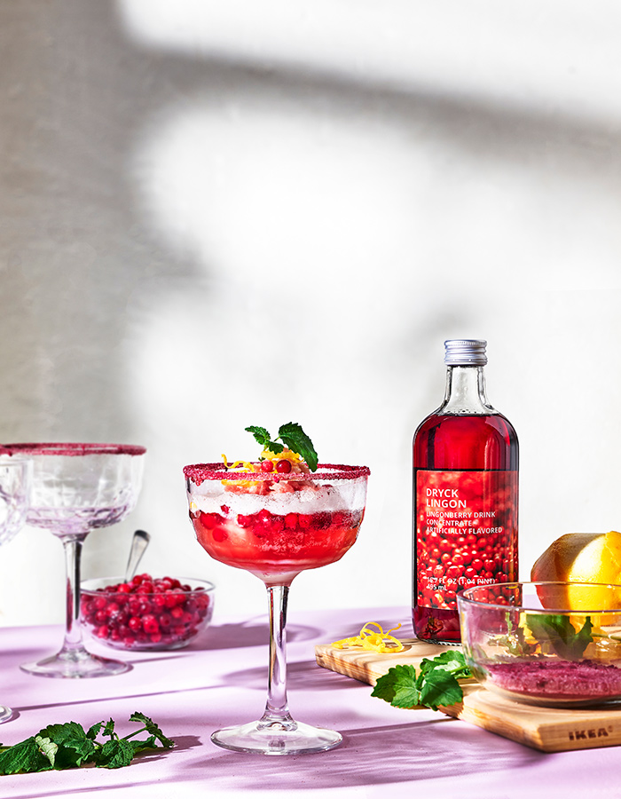A glass of lingonberry punch with a sugared rim and mint garnish sits on a purple surface next to a bottle of DRYCK LINGON concentrate. 