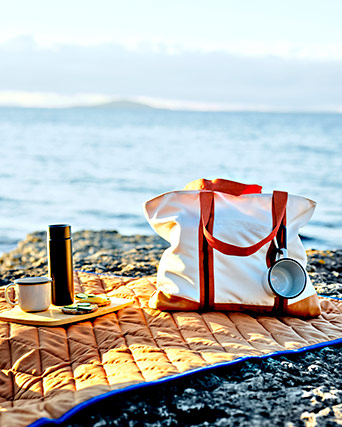 Manta de picnic naranja sobre rocas frente al mar, con una bolsa blanca y naranja, un termo negro, tazas y una tabla con comida.