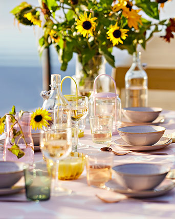 Mesa de exterior decorada con platos lilas, copas de vidrio, farolillos de colores y un gran jarrón con flores amarillas.