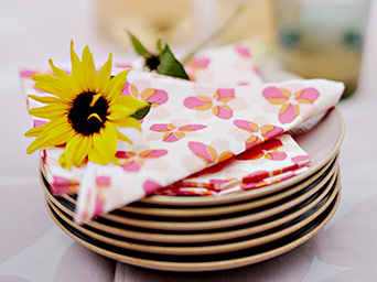 Pila de platos con servilletas de patrón floral rosa y una flor amarilla encima, en un ambiente luminoso.