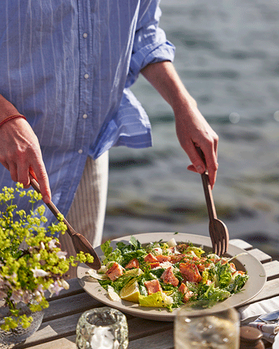 Una fotografía de primer plano de una persona con una camisa a rayas azules sirviendo una ensalada con trozos de salmón de un plato blanco usando cubiertos de madera, en una mesa de madera al aire libre junto al agua.