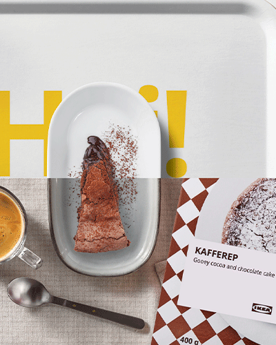 Una fotografía desde arriba de una porción de tarta de chocolate en un plato, una taza de café, una cuchara y un paquete de KAFFEREP de IKEA sobre una servilleta de tela. El fondo muestra parte de una bandeja blanca con el texto "H!" y el logotipo de IKEA.