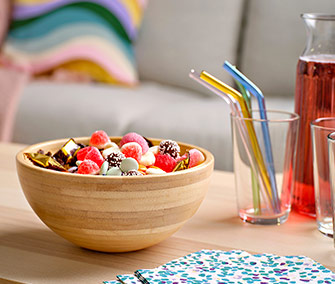 Cuenco de bambú con caramelos variados sobre mesa de madera, junto a vasos con pajitas de colores y una jarra con bebida rosada.