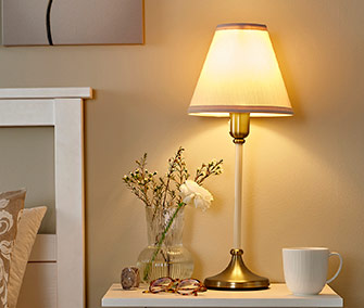 Lámpara de mesa clásica con luz cálida sobre mesita de noche blanca, acompañada de un jarrón con flores pequeñas y una taza blanca.