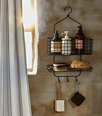 Organizador de ducha colgante en metal negro con botes de jabón, esponjas naturales y accesorios, colgado en una pared de aspecto rústico.