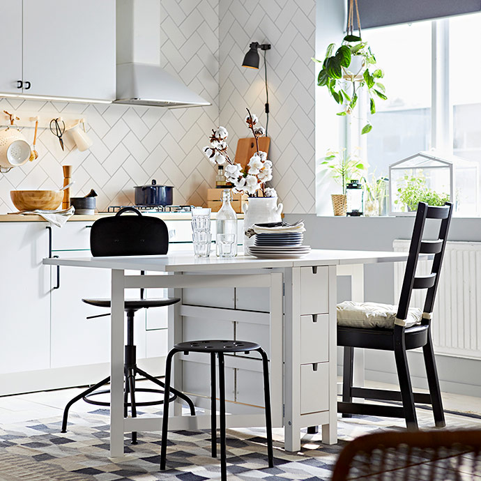 Comedor luminoso con mesa blanca de alas abatibles, sillas negras de distintos estilos y una alfombra de cuadros grises sobre suelo blanco.