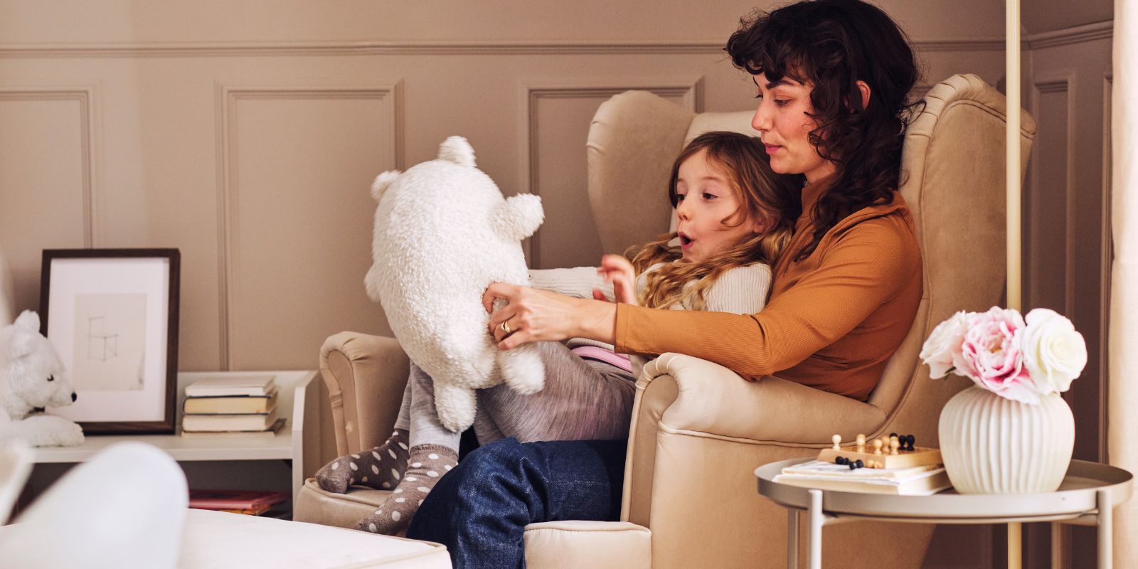 Madre y niña sentadas en un sillón en un salón acogedor, jugando con un peluche junto a una mesa con flores y una bandeja.