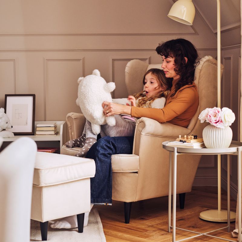 Madre y niña sentadas en un sillón en un salón acogedor, jugando con un peluche junto a una mesa con flores y una bandeja.