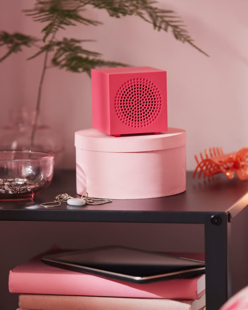 Primer plano de un altavoz Bluetooth portátil KALLSUP de color rosa, colocado sobre una mesita de noche negra junto con algunos objetos decorativos.