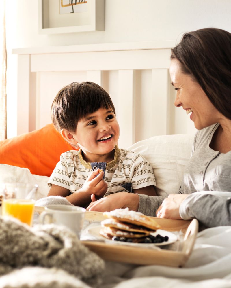 Madre y niño desayunando en la cama, compartiendo un momento alegre con zumo y tortitas en una bandeja.