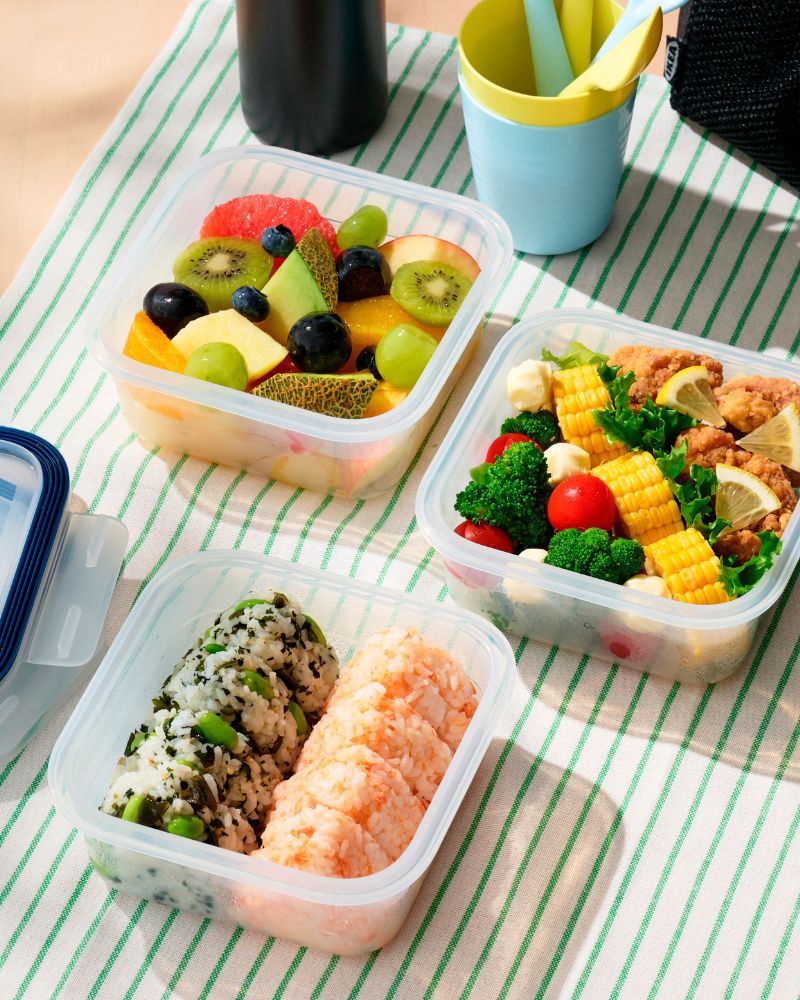 Tres recipientes con onigiri de colores, frutas frescas, brócoli y maíz sobre un mantel de rayas verdes. Una comida fresca y organizada.