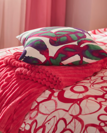 Un primer plano de una funda de cojín multicolor PELARKÖRSBÄR colocada sobre una colcha rosa brillante GULLSTRÅLE.