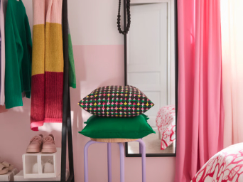 Un dormitorio con una funda de cojín ÄLVSJÖSKOGEN en tonos rosa y verde colocada sobre una funda de cojín GURLI verde, apilada sobre un taburete.
