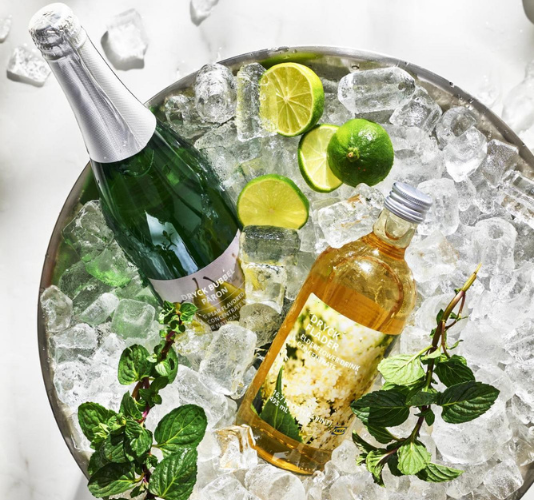 Botellas de concentrado para bebida con lima, menta y hielo en una cubeta metálica llena de hielo.