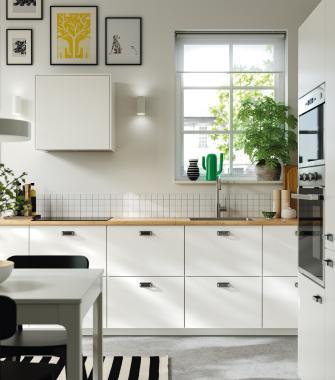 Modern white kitchen with glass-front upper cabinets, a light countertop, plants, and kitchen utensils near a window.