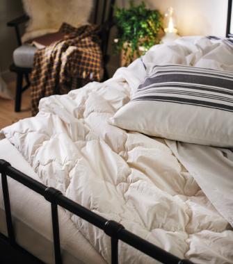 Close-up of an unmade bed featuring a fluffy white duvet, a grey striped pillow, and a black metal bed frame. In the background, a chair with a checkered throw blanket and a green plant are visible under warm lighting.