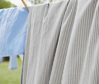 Grey and white striped sheets and a light blue garment hanging on an outdoor clothesline. The background shows a blurred green garden under bright daylight.