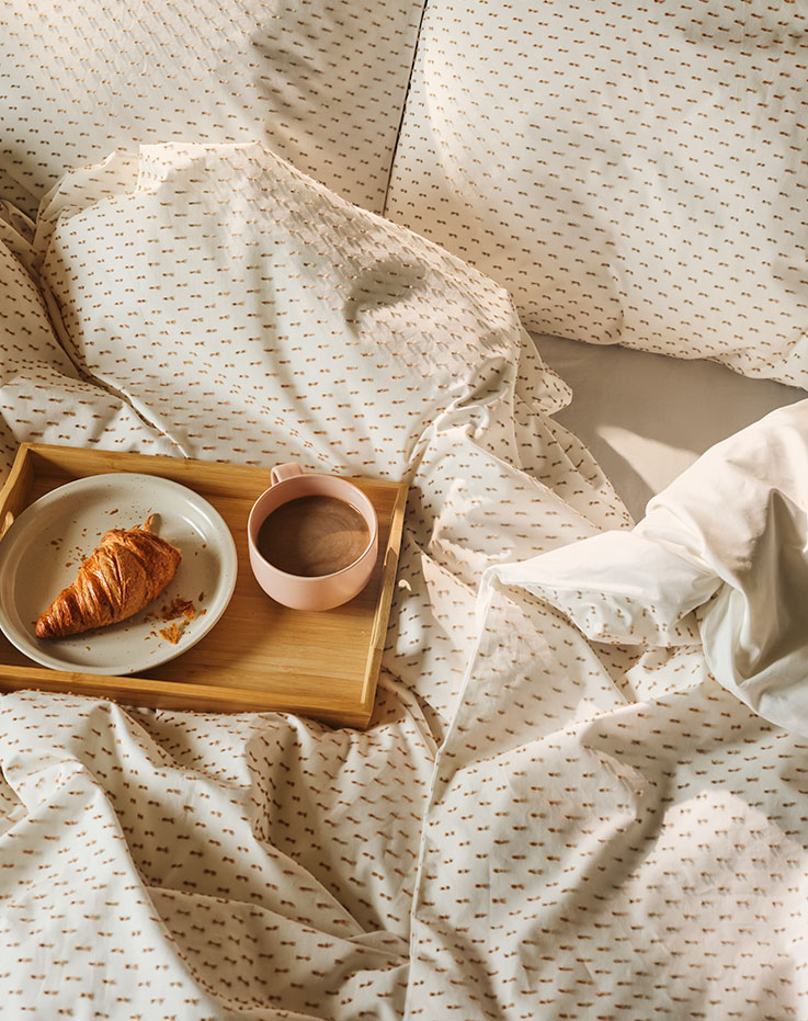A bamboo FRUKTSKAL tray, with a cup of coffee and a croissant on a plate, sits on a bed with BANDKNIPPE bed linen.