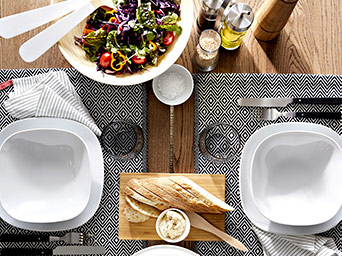 Vista superior de una mesa servida con platos blancos, ensalada colorida, pan artesanal y manteles individuales con patrón geométrico negro.