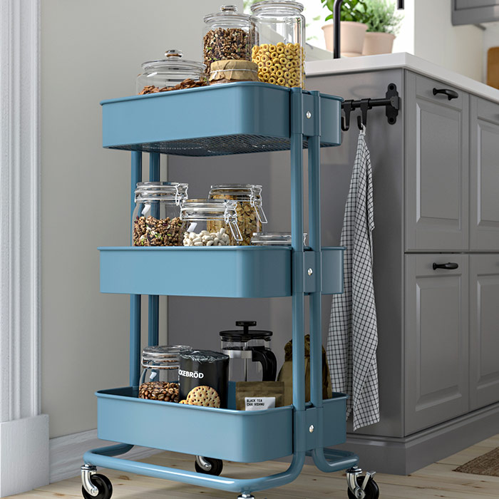 Carrito de cocina azul con tres niveles lleno de frascos de vidrio con alimentos, cereales y café junto a un mueble gris.
