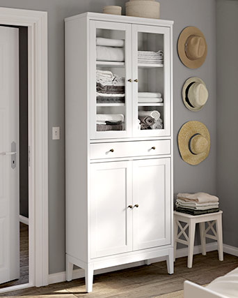 Armario blanco con puertas de vidrio que muestran toallas ordenadas, ubicado junto a una pared gris decorada con sombreros de paja.