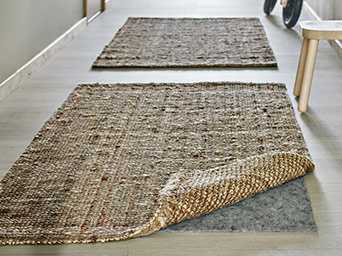 Dos alfombras de fibra natural en un pasillo, con una esquina levantada mostrando una base antideslizante gris sobre el suelo de madera.