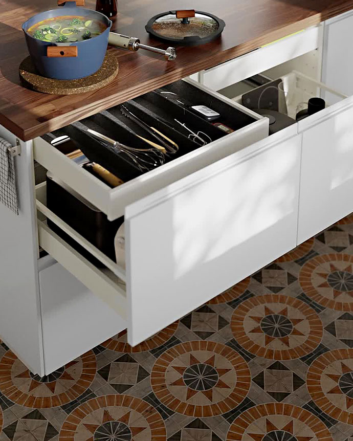 Cajón de cocina blanco abierto con organizador de cubiertos negro sobre un suelo de mosaico geométrico y encimera de madera.