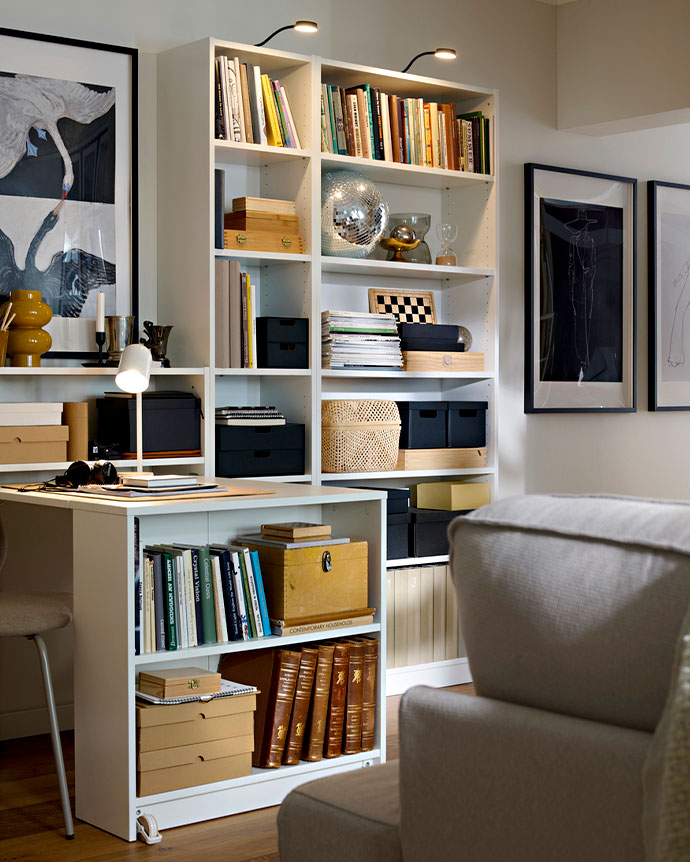 Librería blanca con escritorio integrado, llena de libros, cajas organizadoras, una bola de discoteca y cuadros decorativos en un salón luminoso.