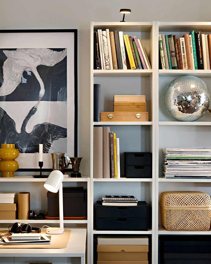 Detalle de estantería blanca con libros, una bola de discoteca, cajas de bambú y un flexo iluminando un escritorio con carpetas y un cuadro de aves.