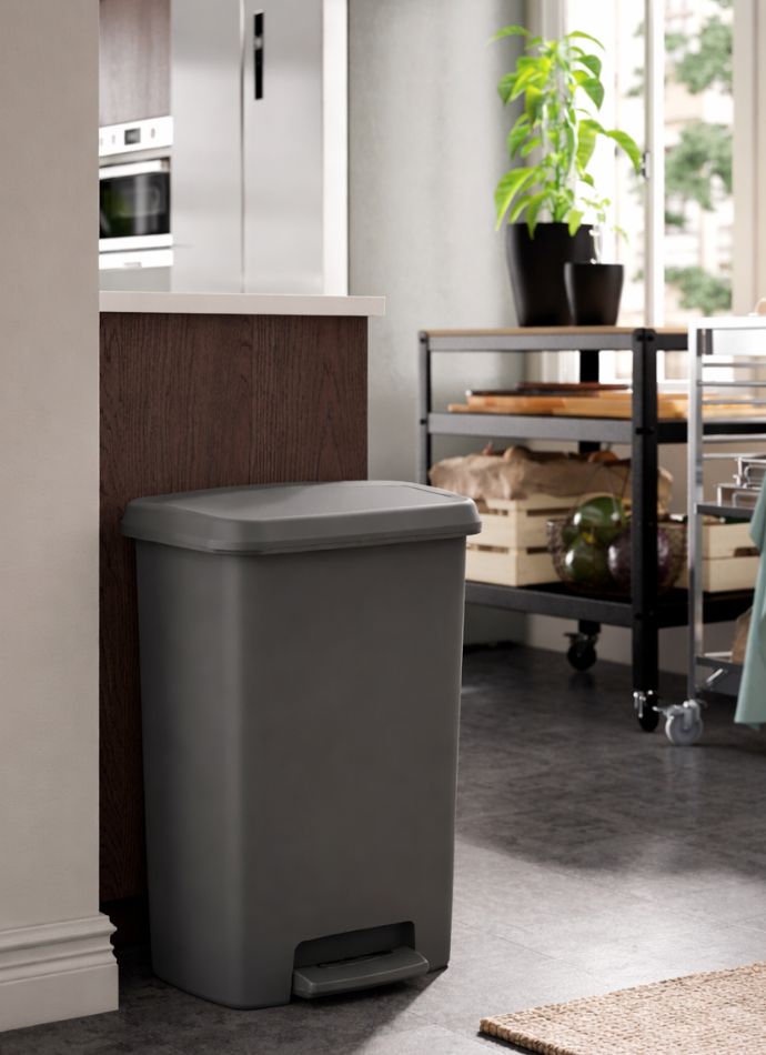 Grey trash can with metallic lid in a modern kitchen with a utility cart and green plant.