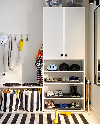 Armario blanco abierto con estantes para cascos y zapatos, junto a una zona de colgar con bolsas de rayas y botas amarillas en un suelo radiado.