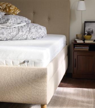 Beige upholstered bed with a white mattress and patterned bedding next to a wooden nightstand with a lamp and decor.