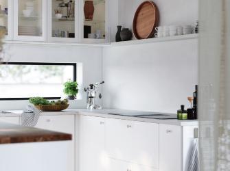 Modern white kitchen with glass-front upper cabinets, a light countertop, plants, and kitchen utensils near a window.