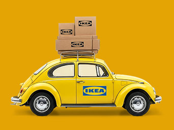 Side profile of a yellow vintage car with the IKEA logo on the door, carrying several cardboard boxes on its roof rack against a yellow background.