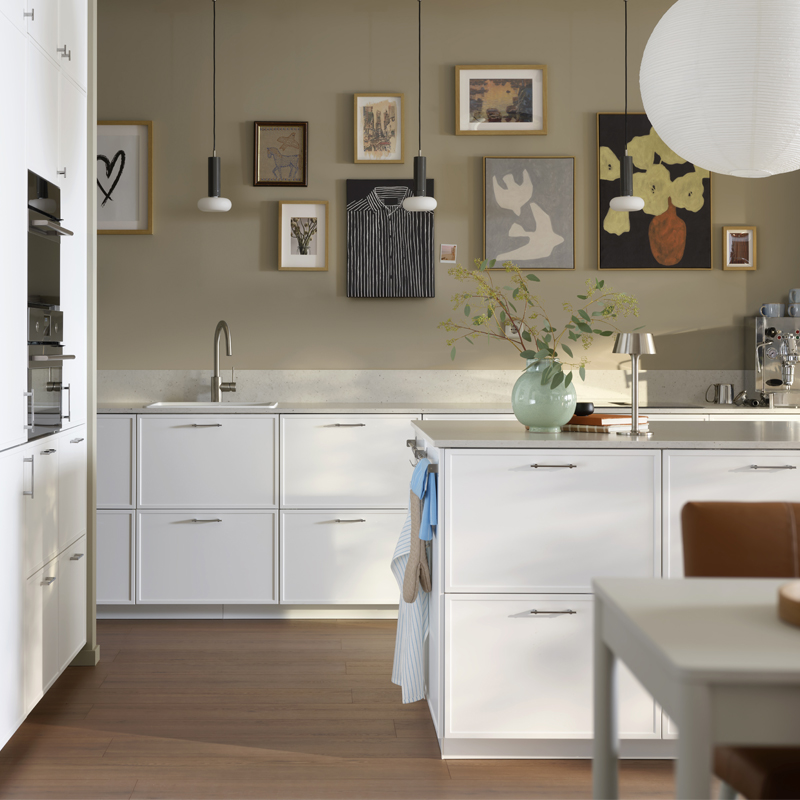 A modern Nordic-style kitchen with white cabinets, a light-colored stone countertop, a wall decorated with a gallery of paintings, and a central island with accessories.
