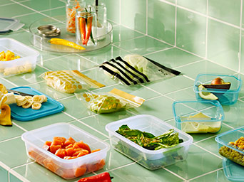 Varios recipientes transparentes y bolsas de plástico con dibujos llenos de ingredientes frescos sobre una encimera de azulejos verdes.