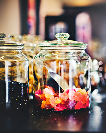 Primer plano de botes de vidrio con tapa llenos de caramelos de colores en forma de corazón y granos oscuros sobre una superficie oscura.