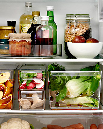 Interior de nevera organizado con botes de vidrio, botellas y recipientes apilables con fruta fresca, rábanos y verduras de hoja verde.