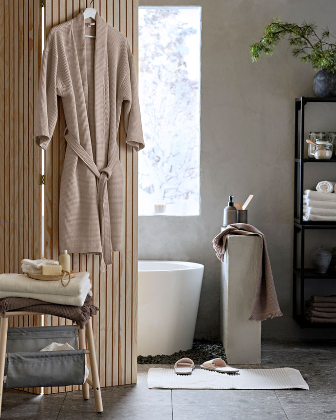 Un albornoz beige cuelga de un biombo de madera en un baño luminoso con bañera blanca, toallas ordenadas, zapatillas y una atmósfera relajante.