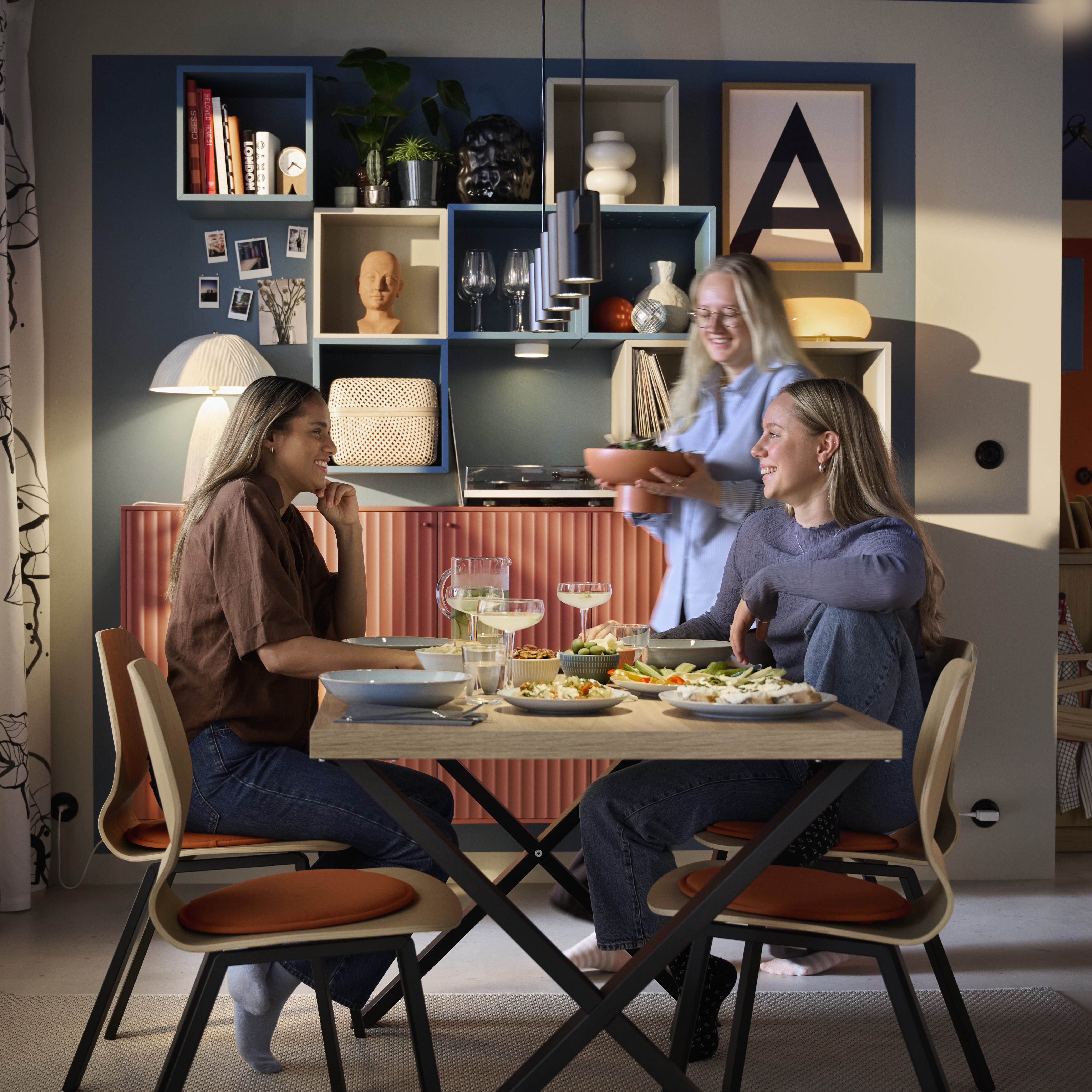 Three friends having dinner in a modern dining area with colorful shelving and warm, cozy lighting.