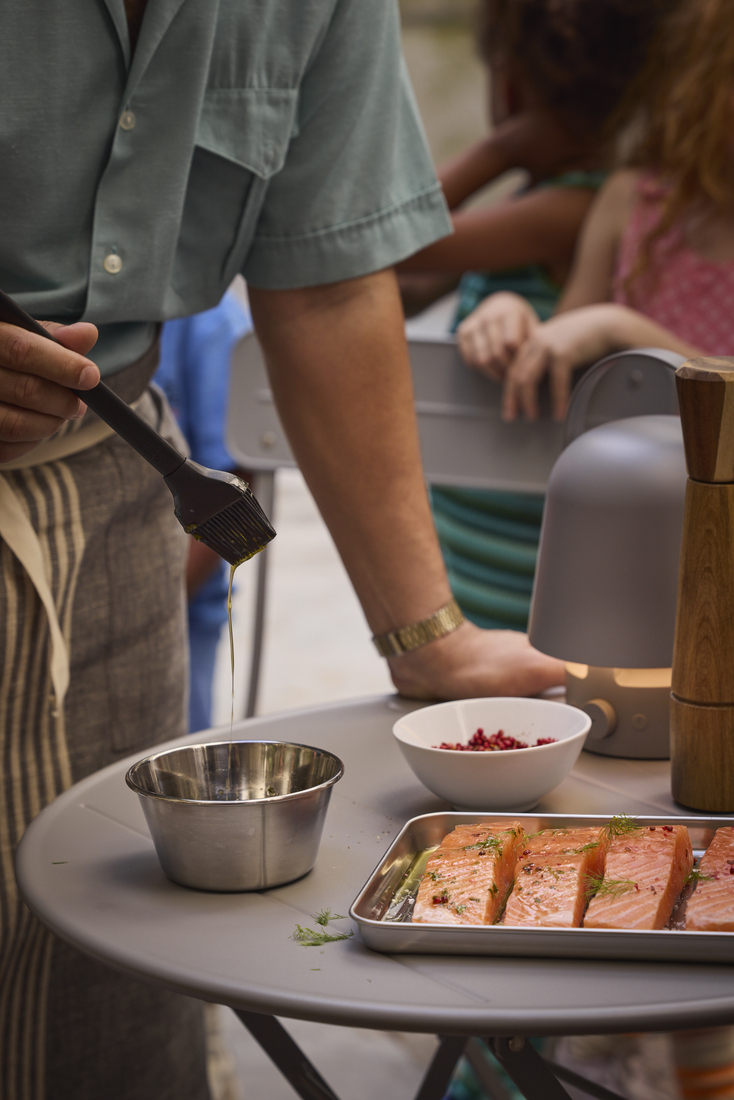 Close-up of a man brushing glaze onto raw salmon fillets on an outdoor table next to bowls of spices.